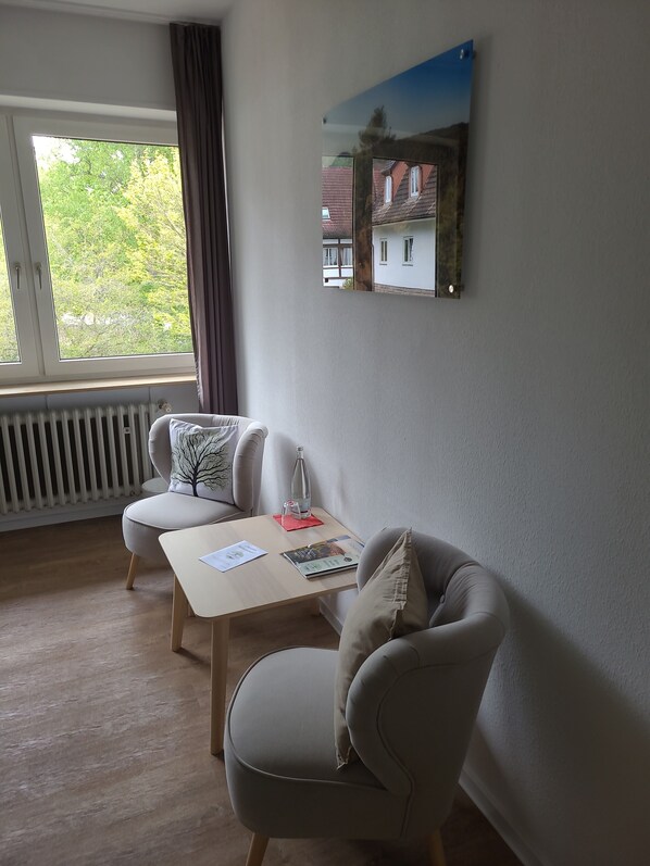 A modern living room with a sofa, armchair, and a small table with a bottle and papers. A window with curtains is visible, and a framed picture hangs on the wall. - Die Heilsbach (Schönau)