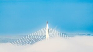 A bridge with a central tower and cables extending outward, surrounded by clouds.