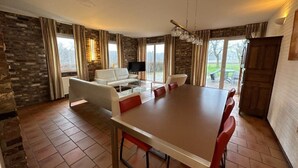Un espace repas comprenant une grande table et des chaises, un canapé et un téléviseur, le tout dans une pièce aux murs en pierre et aux grandes fenêtres.