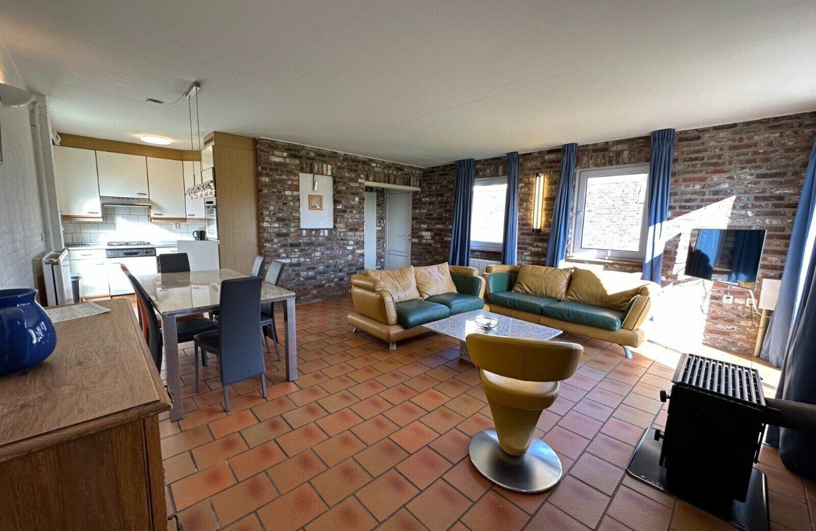 A spacious living area with a sofa set, a dining table, and a kitchen in the background.