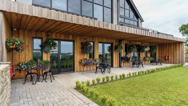A modern building with a wooden facade, large windows, and outdoor seating area with chairs and tables. - The Old House 1147 (Maesteg)