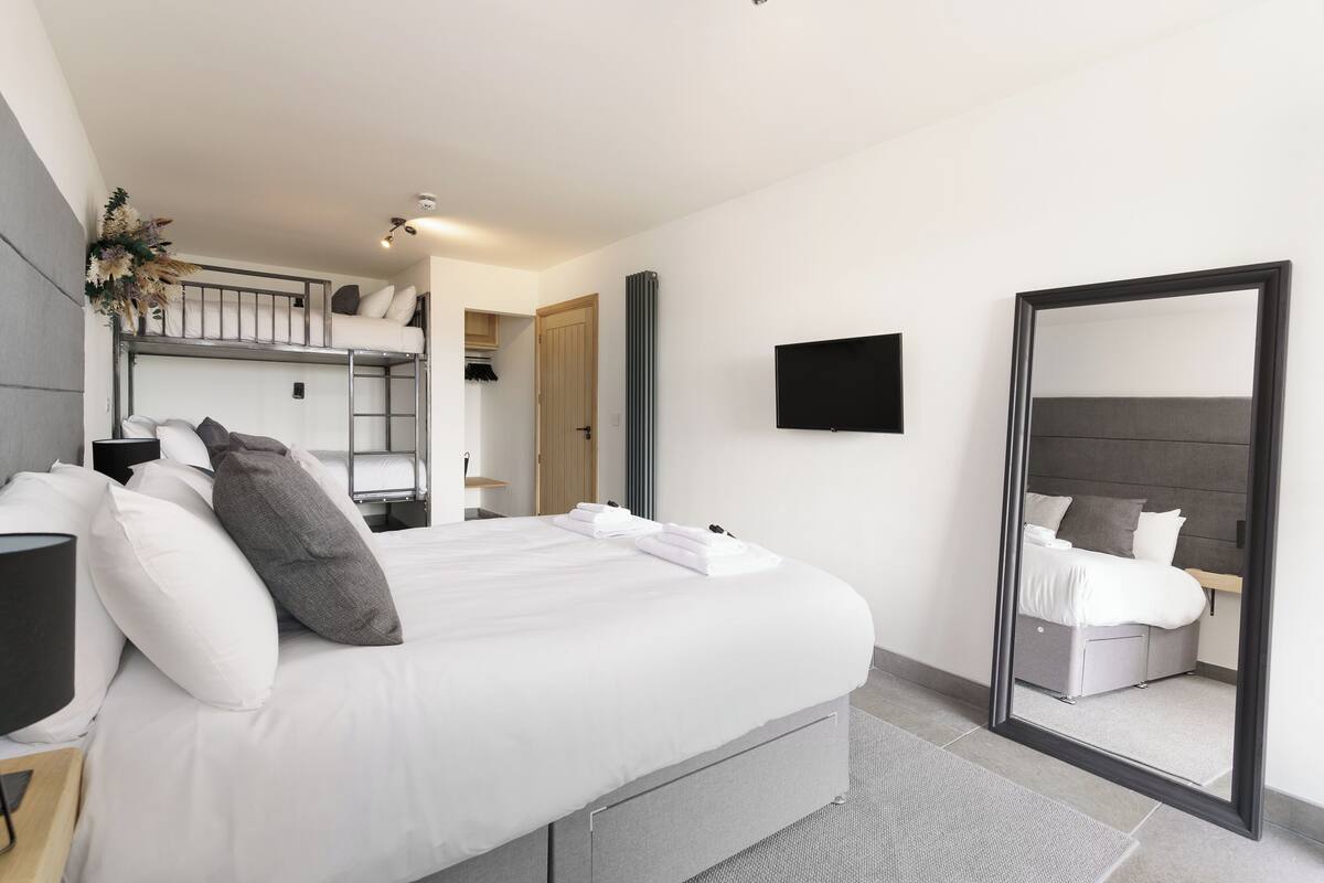 A modern hotel room with a large bed, a mirror, a TV, and a bunk bed in the background.