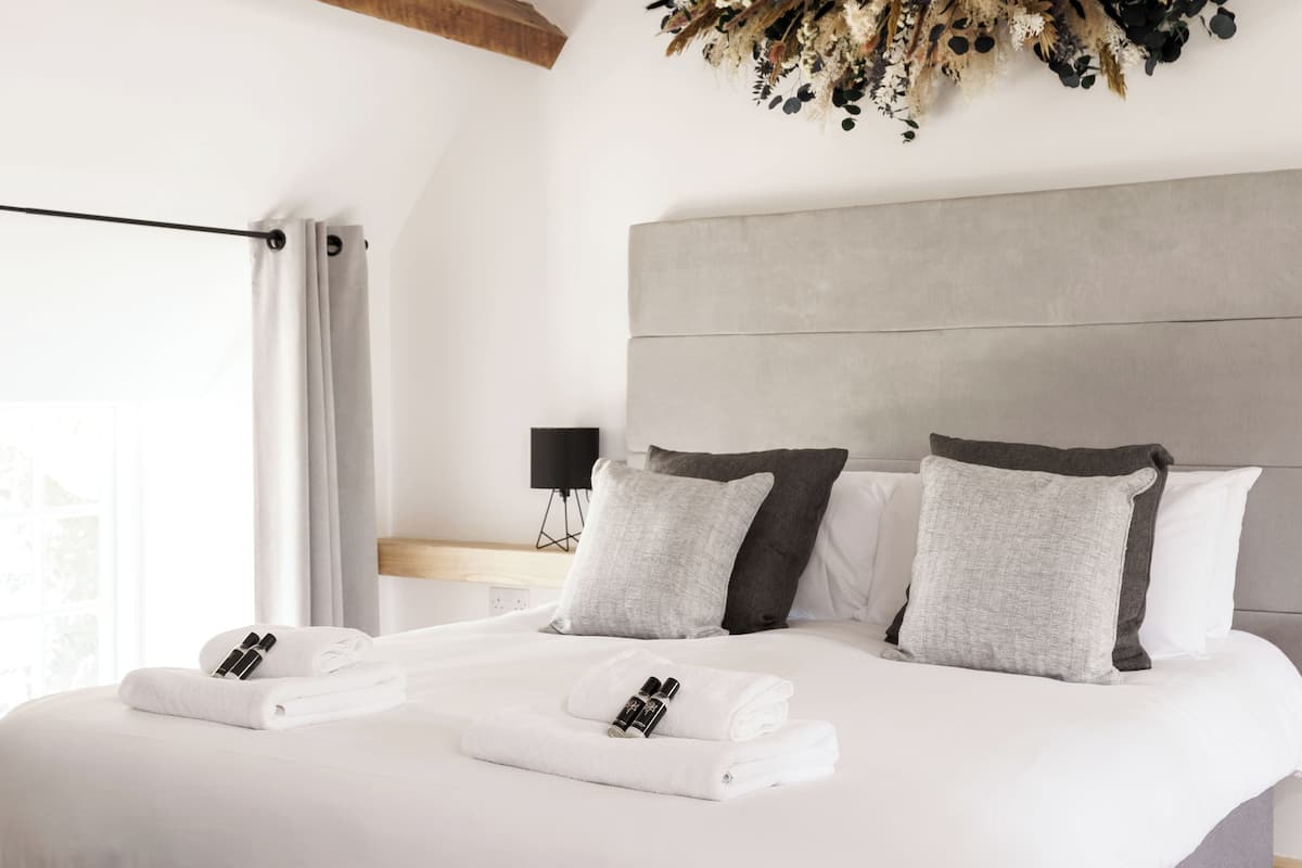 A neatly made bed with white linens and two pillows, a bedside table with a lamp, and a view of the outdoors through a window with curtains.