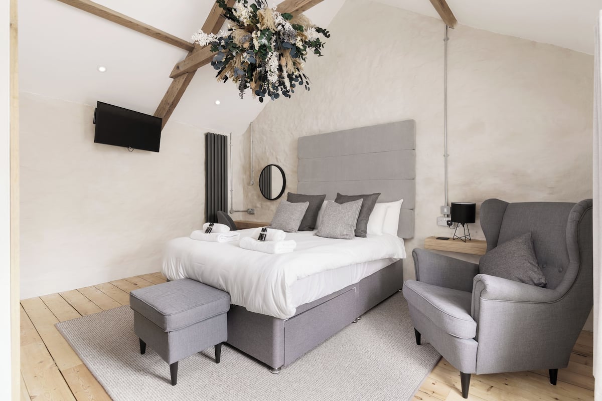 A modern bedroom with a grey bed, matching armchair, and ottoman, a wooden ceiling, and a mounted TV.