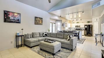 Amplia sala de estar con sofá seccional, reposapiés y una mesa de centro. Cuenta con una zona de cocina con electrodomésticos modernos y una mesa de comedor.