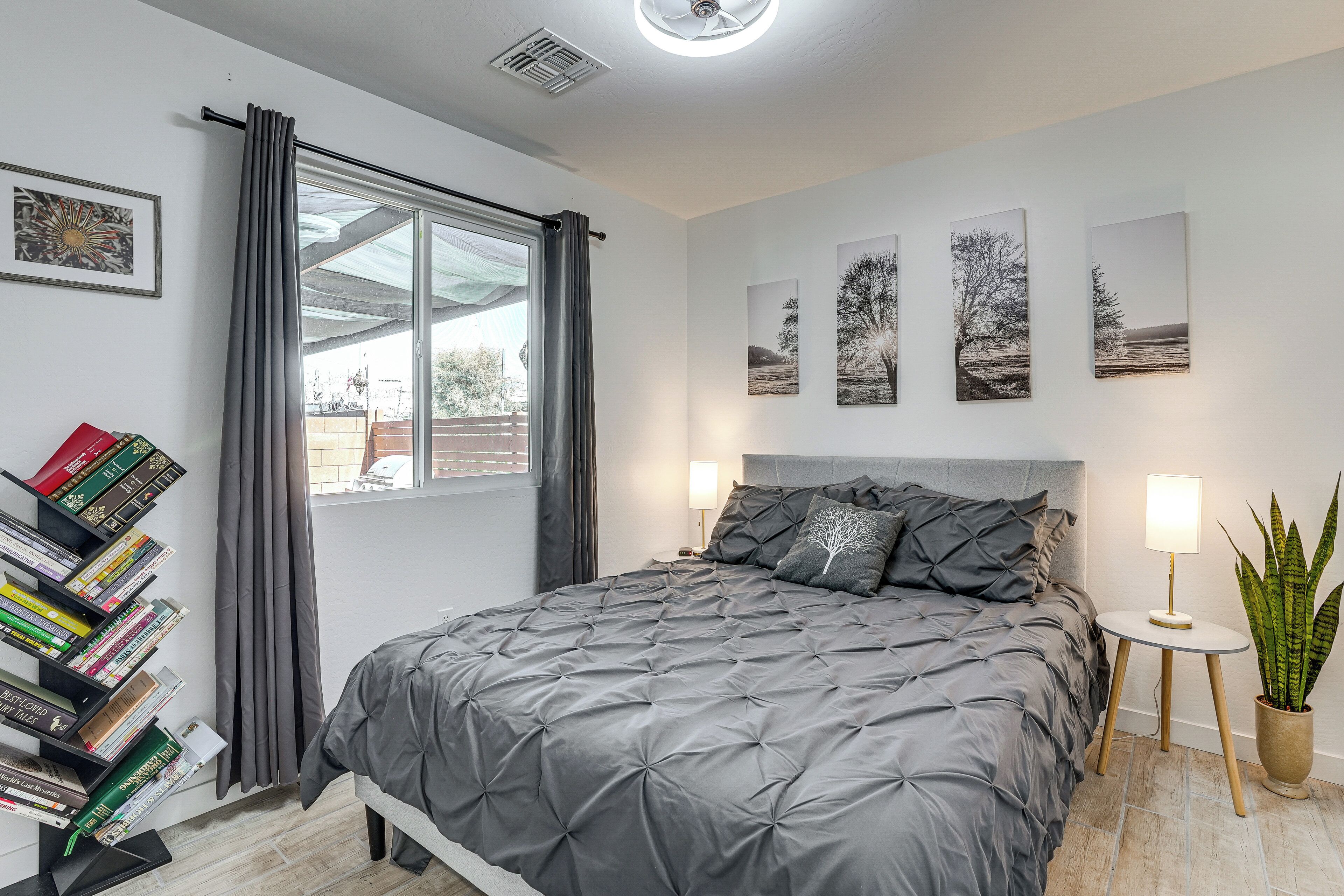 A modern bedroom with a large bed, a bedside table with lamps, a bookshelf, and a window with curtains.