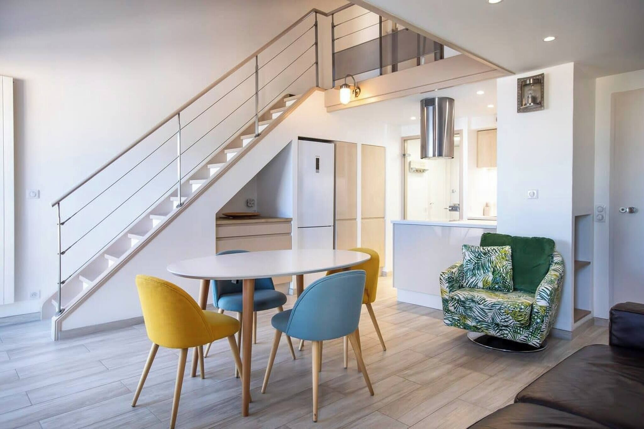 Een moderne leefruimte met een trap, een eethoek met een ronde tafel en stoelen, een keuken en een groene fauteuil met een patroon.