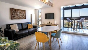 Una sala de estar moderna con un sofá de cuero negro, una mesa de comedor redonda con cuatro sillas, un televisor de pantalla plana montado en la pared y un ventanal con vistas a la ciudad.