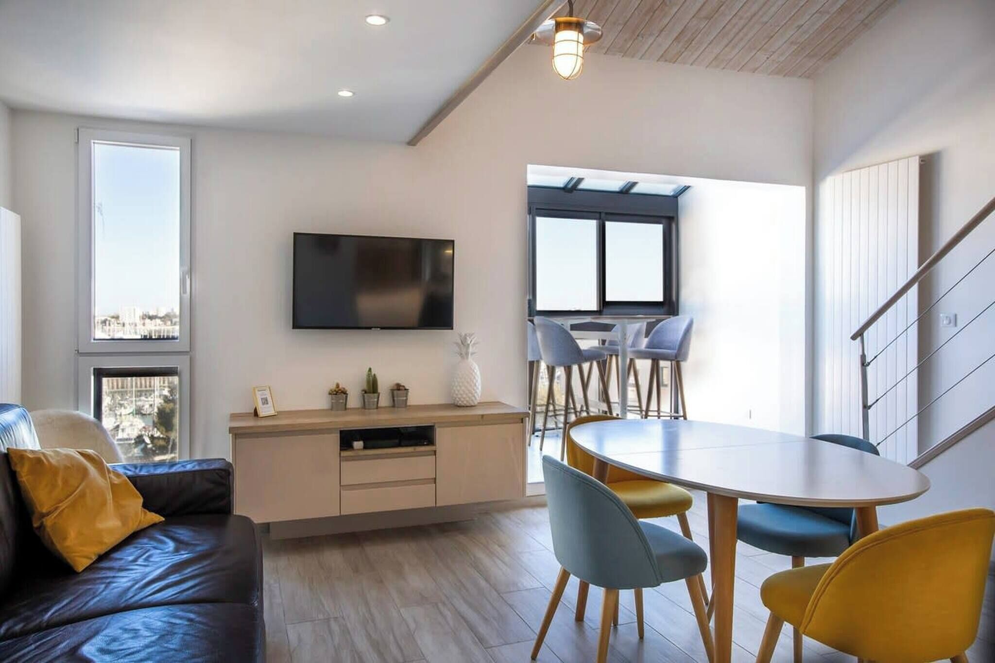 Ein modernes Wohnzimmer mit einer Couch, einem Esstisch mit Stühlen, einem an der Wand befestigten Fernseher und einem Fenster mit Stadtblick.