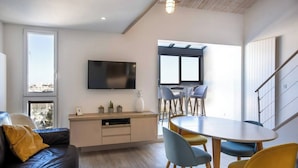 Una sala de estar moderna con un sofá, una mesa de comedor con sillas, un televisor montado en la pared y un ventanal con vistas a la ciudad.