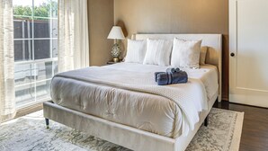A bedroom with a large bed, a nightstand with a lamp, and a view through a window. - 1 Mi to Smu: Home w/ Outdoor Entertainment (Dallas)