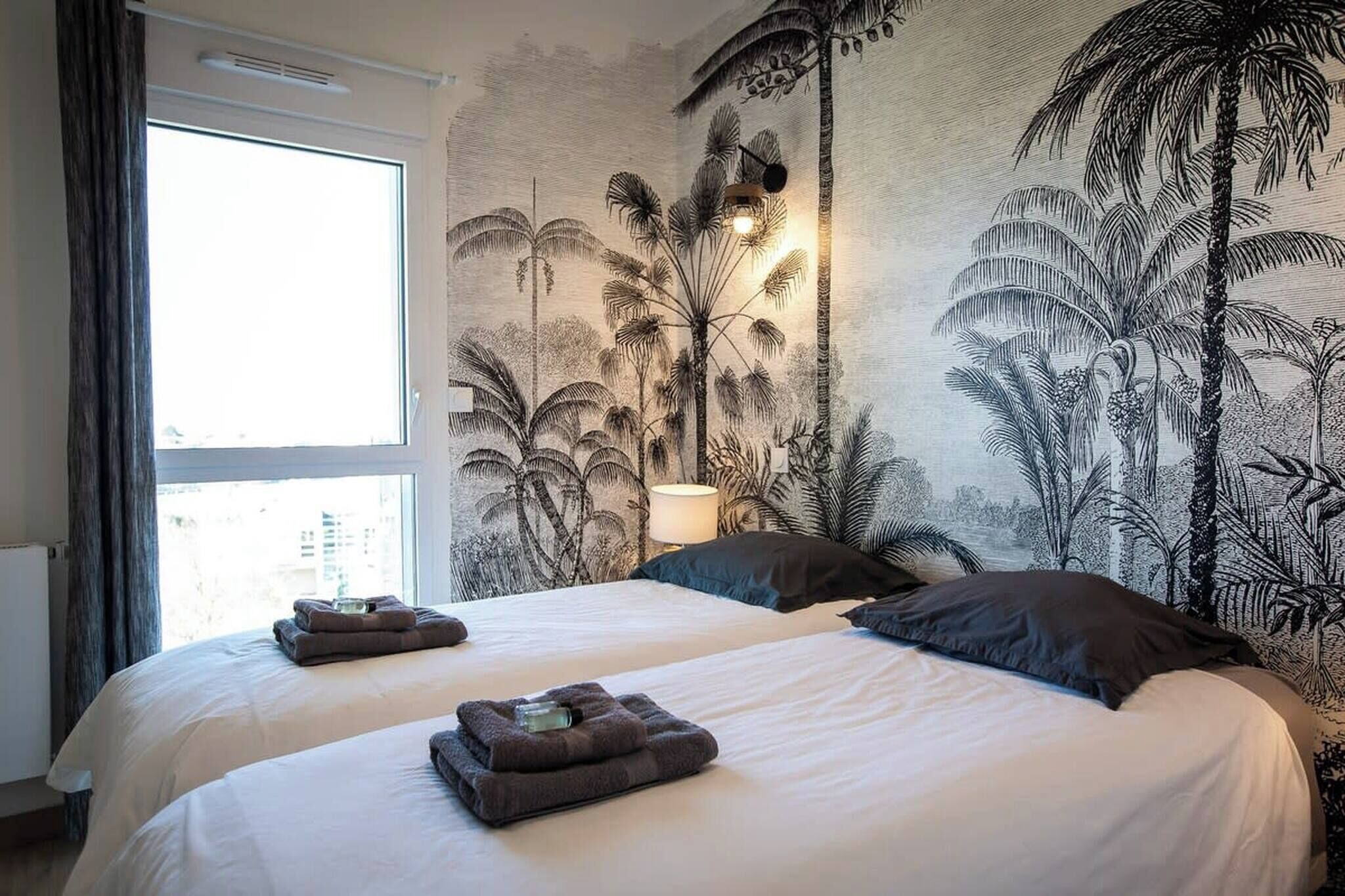 Ein Hotelzimmer mit zwei Betten, einem großen Fenster und einem Wandgemälde von tropischer Vegetation.