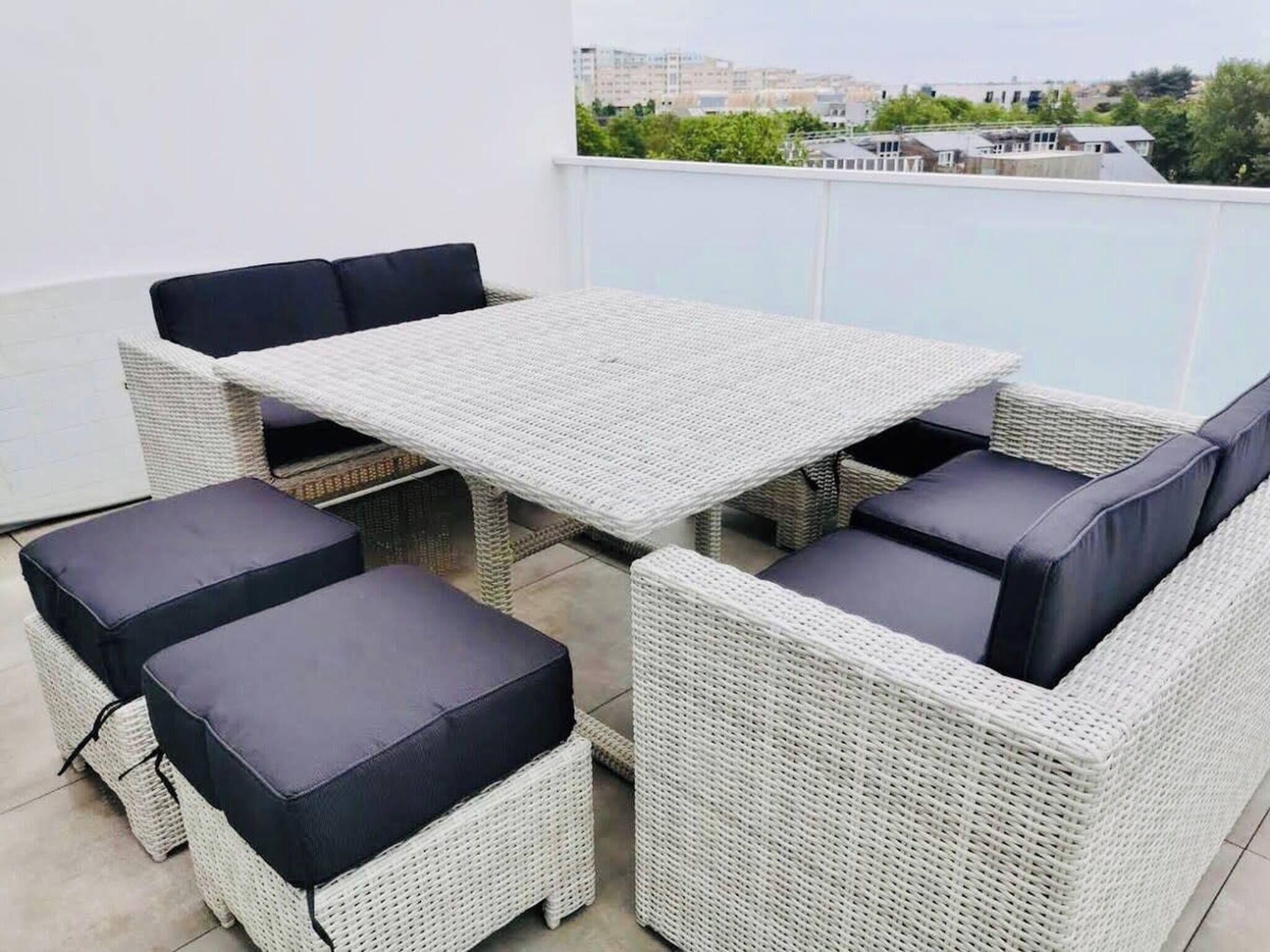 Ein Sitzbereich im Freien mit Korbmöbeln, einem Tisch und gepolsterten Stühlen, vor einer weißen Wand mit Blick auf die Stadt.