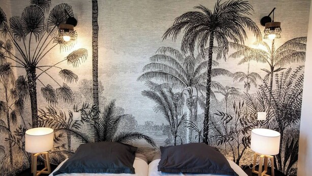 A room with a bed, two bedside lamps, and a wall with a tropical plant mural.