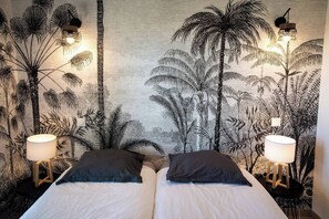 A room with a bed, two bedside lamps, and a wall with a tropical plant mural.