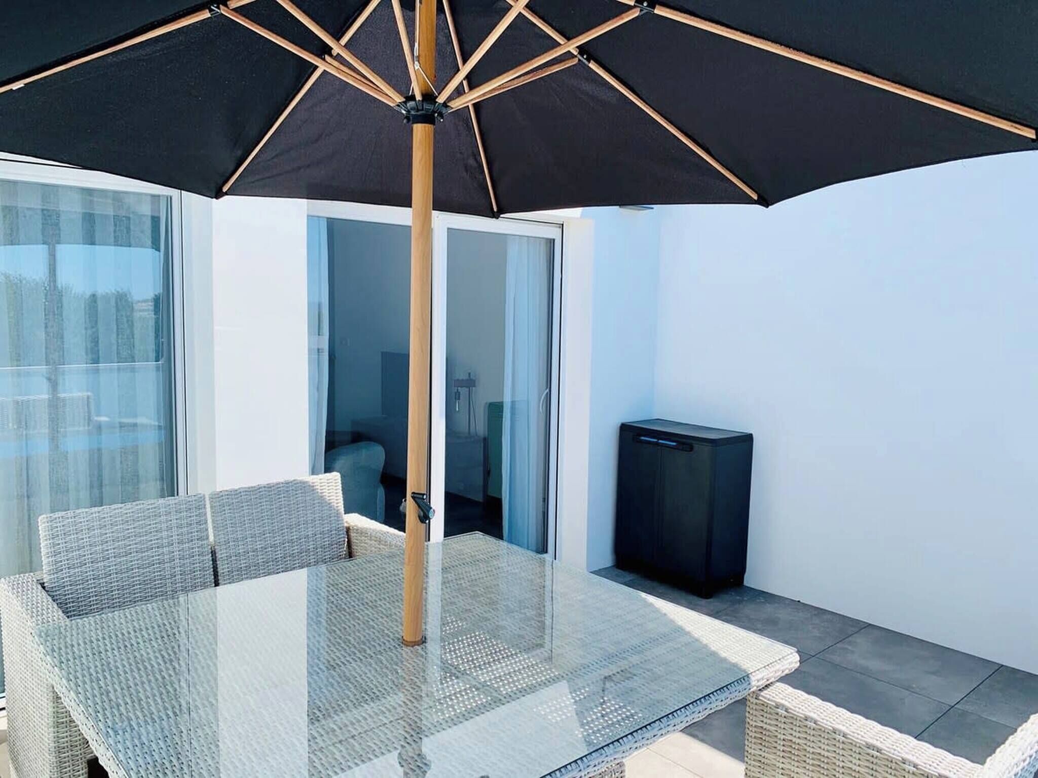 Un espace extérieur moderne, aménagé avec une table et des chaises en verre, un parasol noir et un téléviseur.