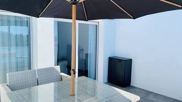 Un espace extérieur moderne, aménagé avec une table et des chaises en verre, un parasol noir et un téléviseur.