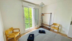 A bedroom with a bed, a wooden chair, a hanging chair, and a balcony with a view of greenery.