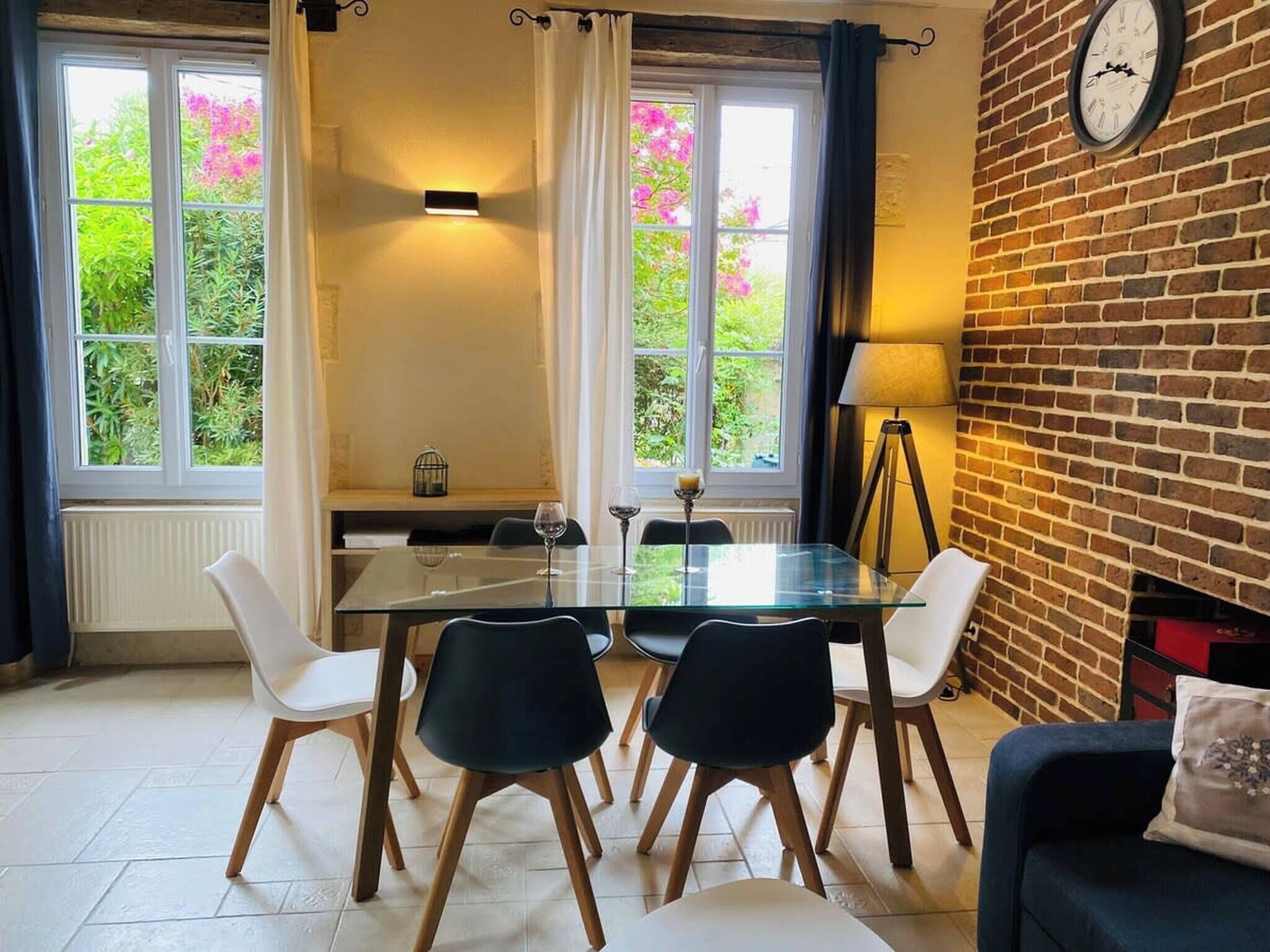 A dining area with a glass table, four chairs, a clock on the wall, and a view of greenery through the windows.