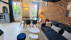 A cozy living room with a black sofa, a glass coffee table, and a dining area with a table and chairs. There is a brick wall with a clock and a window with curtains.