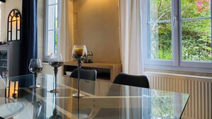 A dining area with a glass table, chairs, and wine glasses. There is a window with curtains and a view of greenery outside.