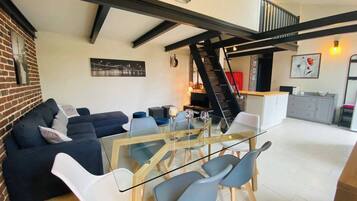 Ein moderner Wohnraum mit einem Glastisch, blauen und weißen Stühlen, einem dunklen Sofa und einer Treppe, die in ein Obergeschoss führt.