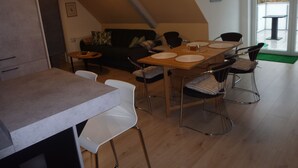 A dining area with a wooden table set for four, surrounded by metal chairs. A dark green sofa is against the wall, and a small round table with a single chair is in the corner.