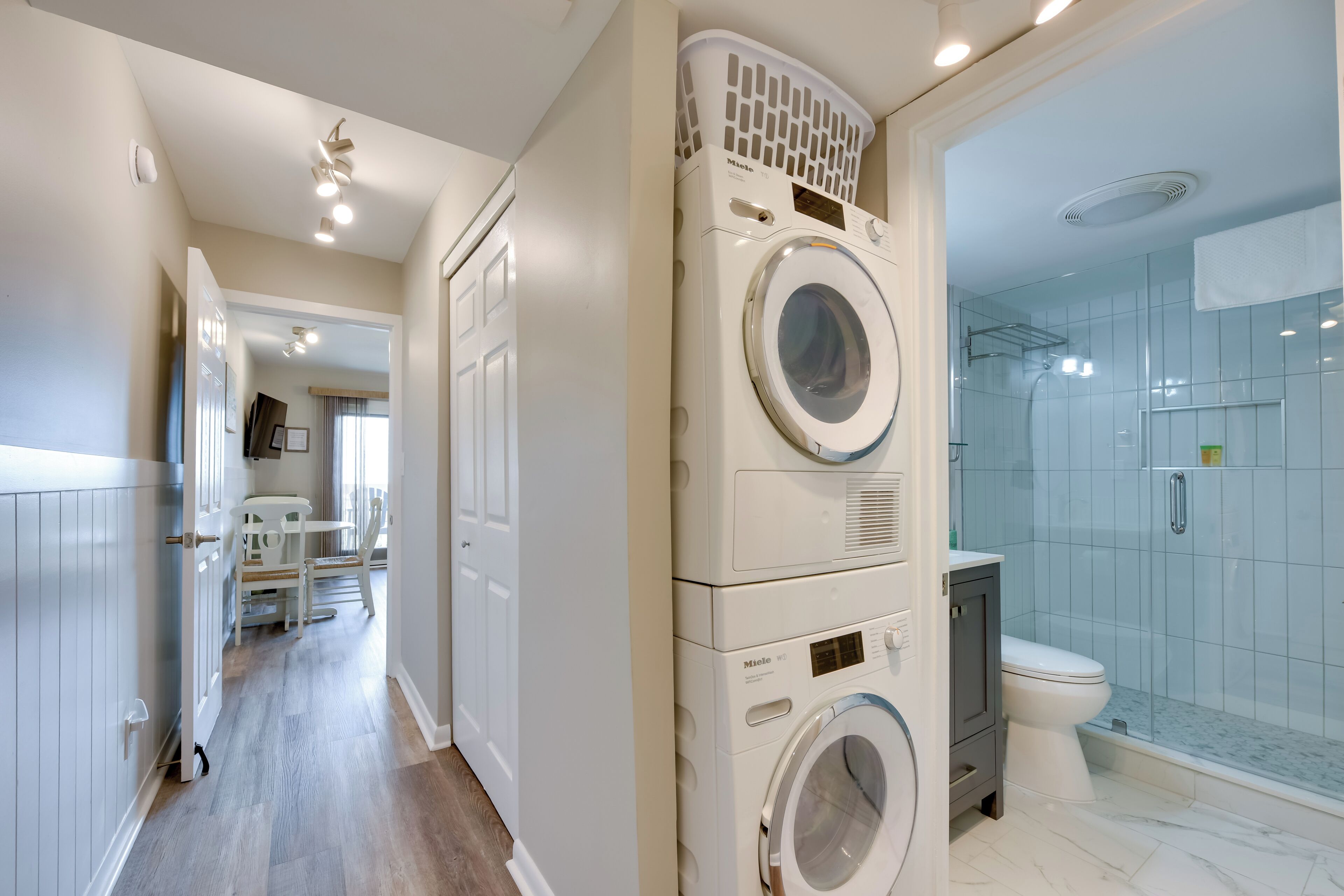 A laundry room with stacked washing machines and dryers, a shower area with glass doors, and a hallway leading to other rooms.