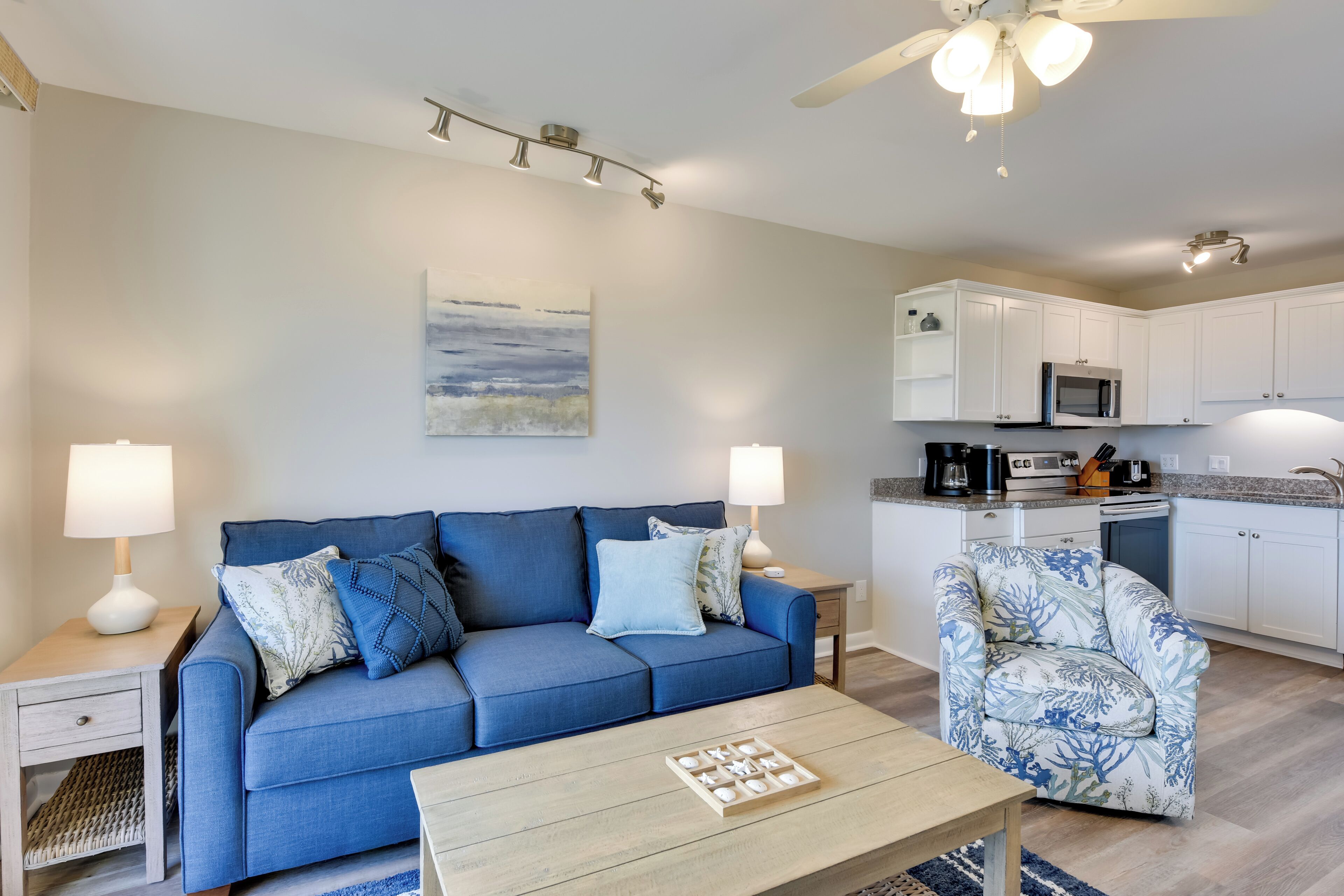 A living room with a blue sofa, a patterned armchair, a coffee table, and a kitchen area with white cabinets and appliances.