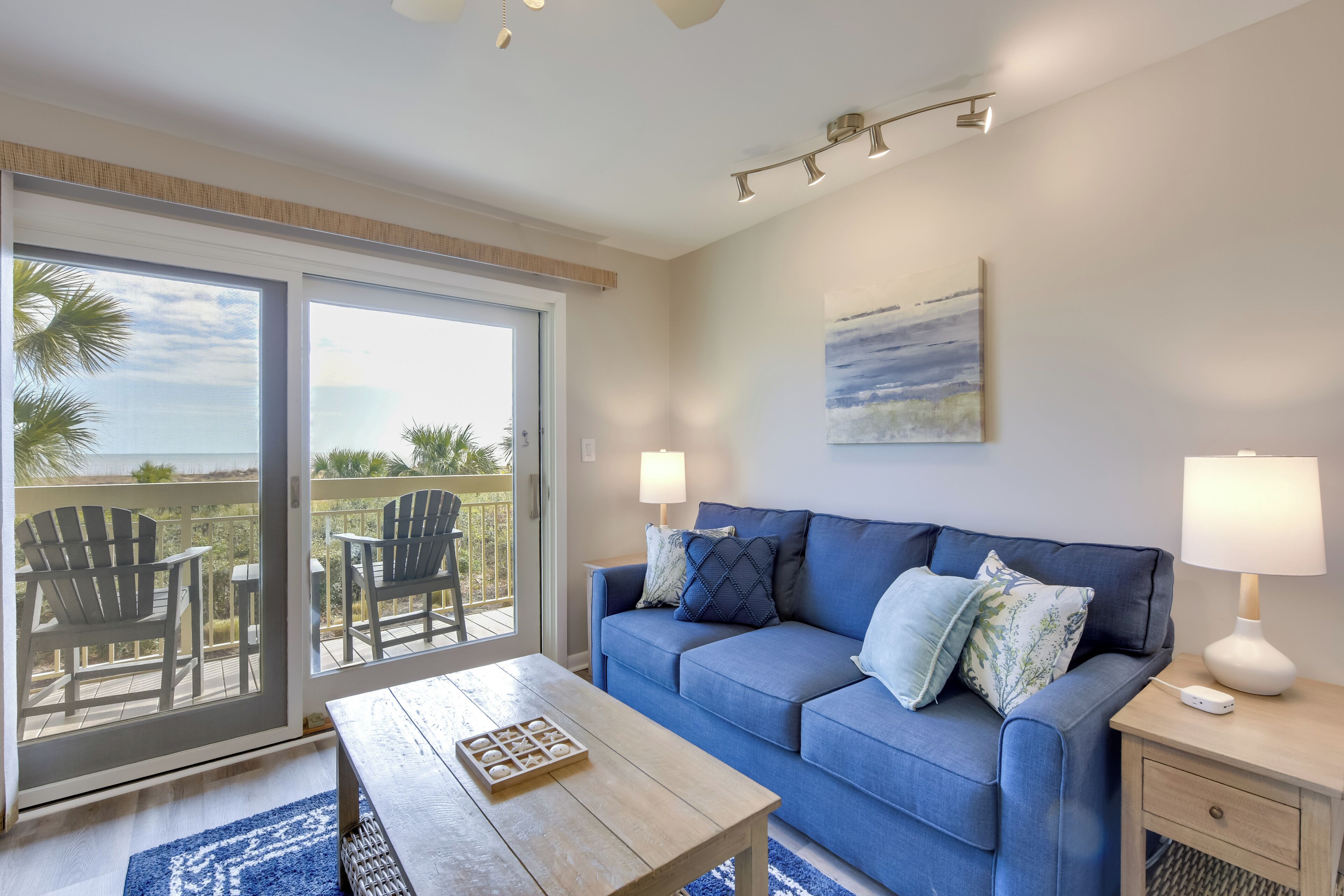 Uma sala de estar moderna com um sofá azul, duas mesas laterais com abajures, uma mesa de centro e uma grande porta de vidro deslizante que dá para uma varanda com móveis de exterior.