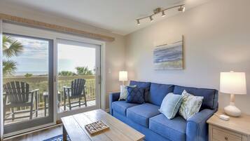 Uma sala de estar moderna com um sofá azul, duas mesas laterais com abajures, uma mesa de centro e uma grande porta de vidro deslizante que dá para uma varanda com móveis de exterior.