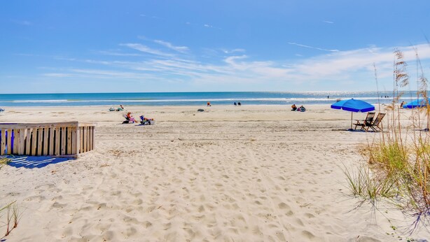 Una playa arenosa con una estructura de madera, sombrillas azules y cielo despejado.