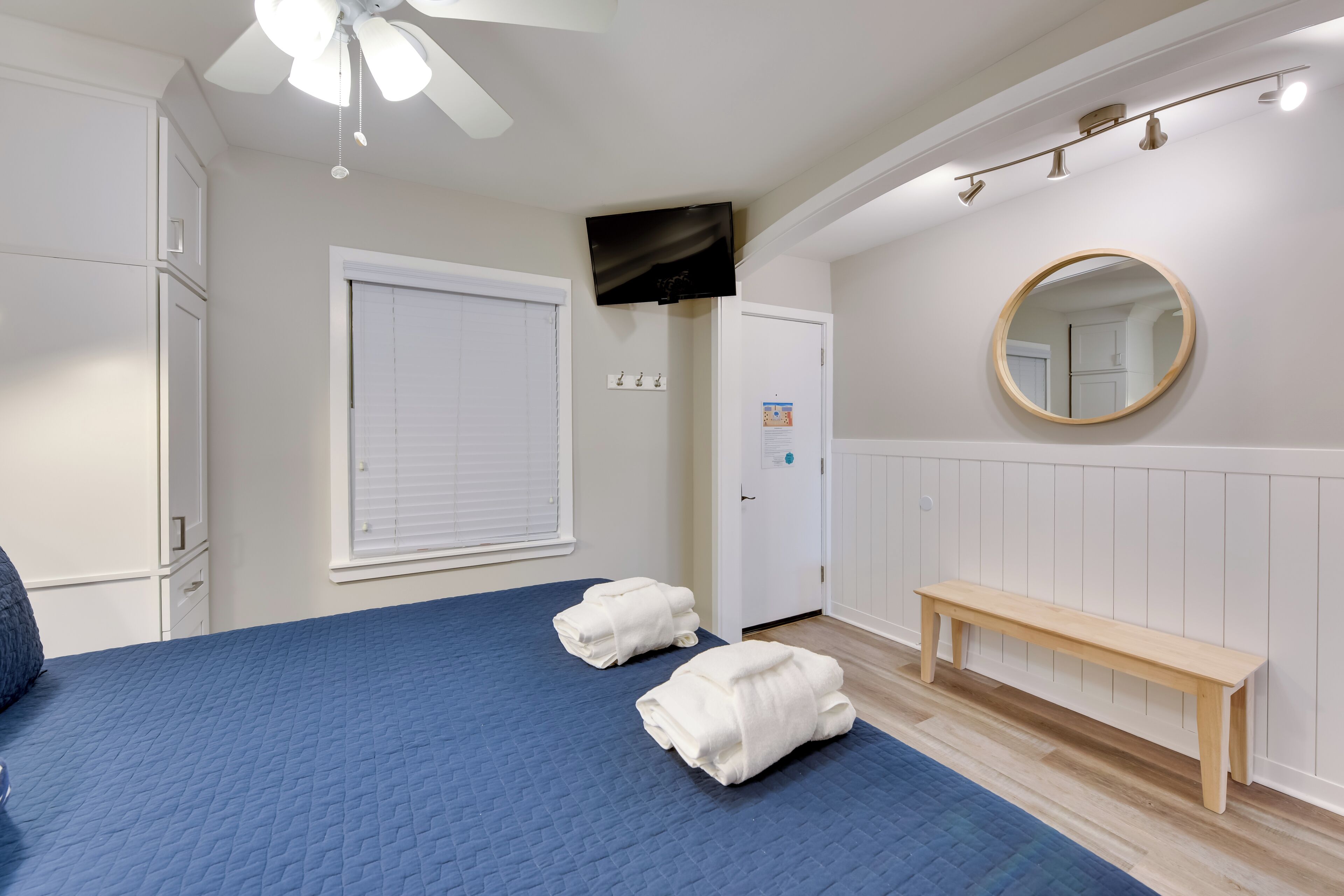 A bedroom with a bed, two towels, a bench, a ceiling fan, a TV, and a mirror.
