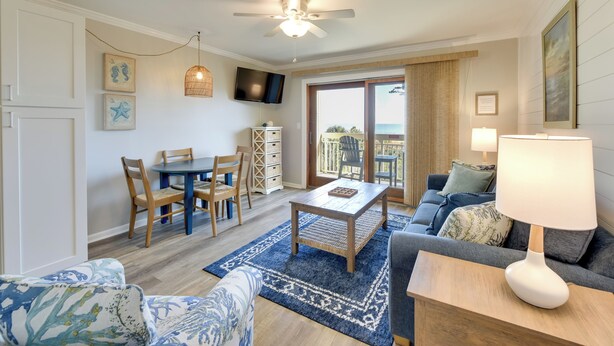 A spacious living area with a ceiling fan, a dining table with chairs, a sofa, a coffee table, and a TV mounted on the wall. - Oceanfront Condo! Breakers 210 w/ Sunrise Views (Hilton Head Island)