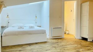 Un grand lit avec du linge de lit blanc, une table de chevet et une lampe. La pièce est dotée d’un plancher en bois et d’une armoire blanche.