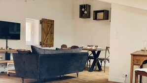 Un salon moderne avec un canapé foncé, une table à manger avec des chaises, une porte en bois et un téléviseur posé sur un meuble.