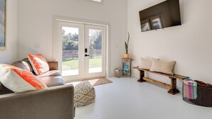 A modern living room with a sofa, a bench, a TV mounted on the wall, and a sliding glass door leading to a garden. - Fish, Boat & Unwind: Gem 6 Mi to Gulf Shores Beach (Gulf Shores)
