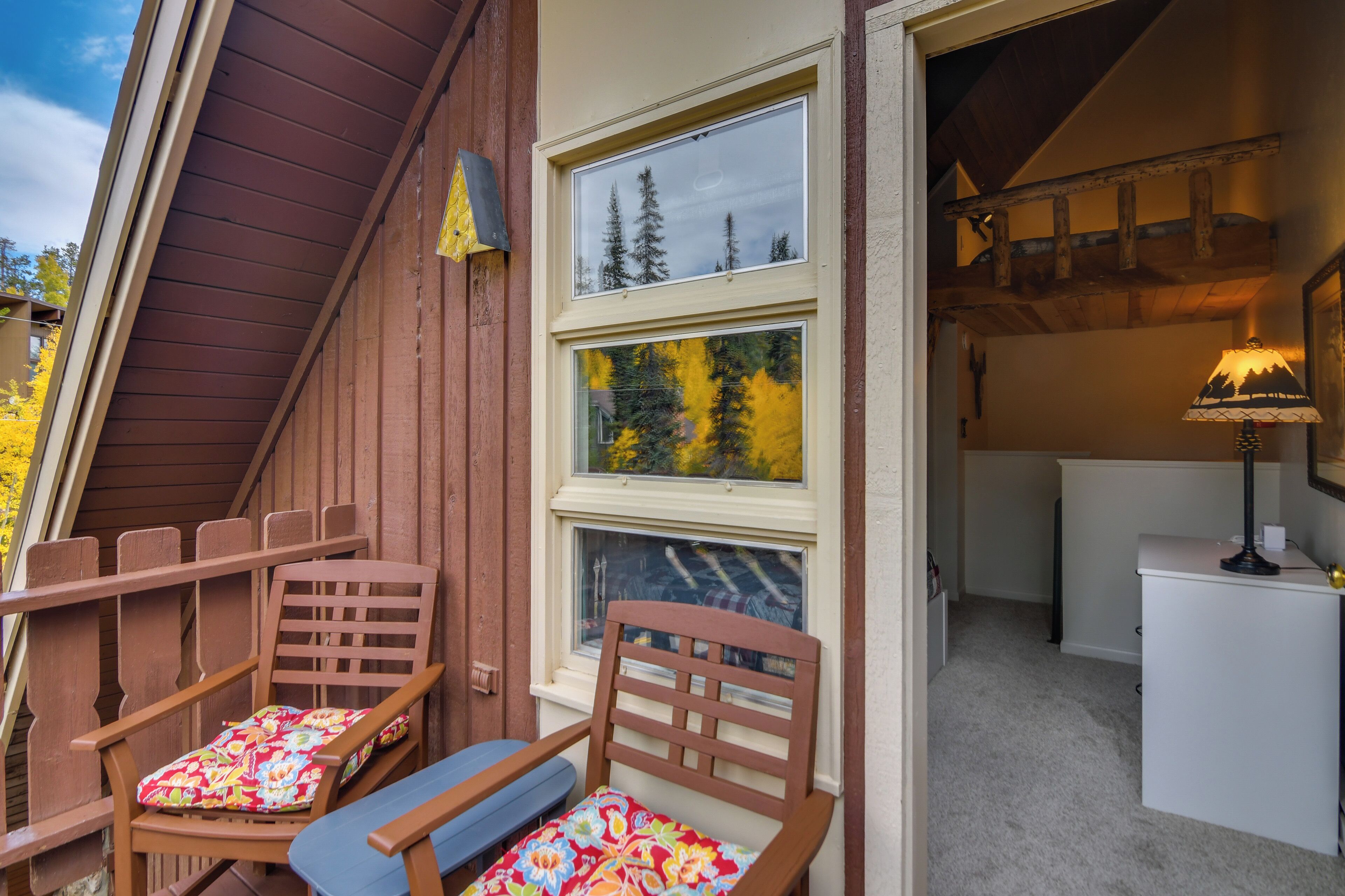 A room with a wooden sloped ceiling, a window with a view of trees, a desk with a lamp, and two chairs with cushions.