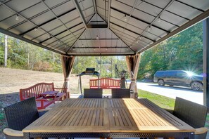 An outdoor covered patio with a wooden table and chairs, a grill, and a view of a road and trees. - Walk to Dollywood! Cabin w/ Pool, Arcade, Gym (Pigeon Forge)