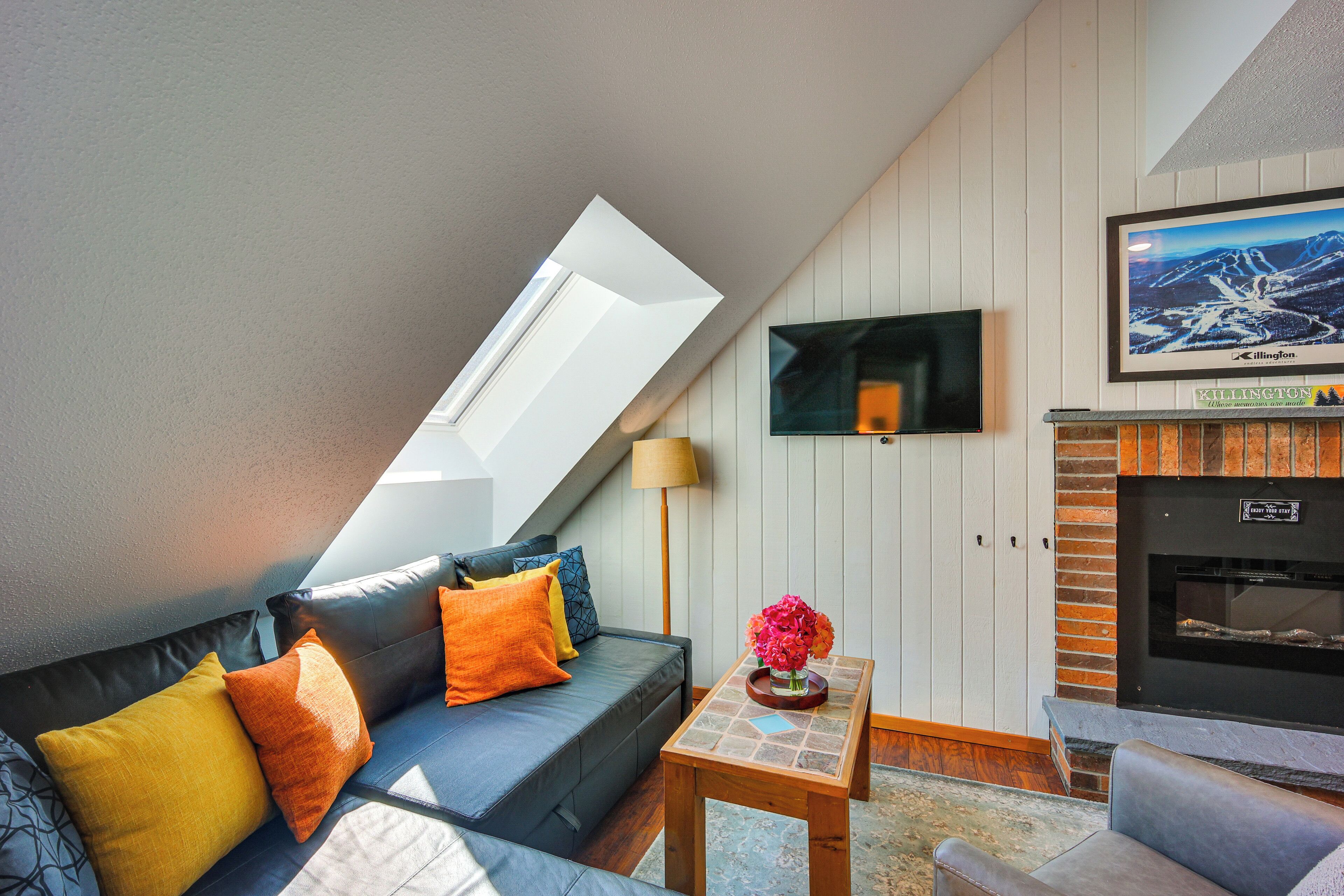 A modern living room with a sloped ceiling, a flat-screen TV mounted on the wall, a fireplace, and a sofa with cushions.