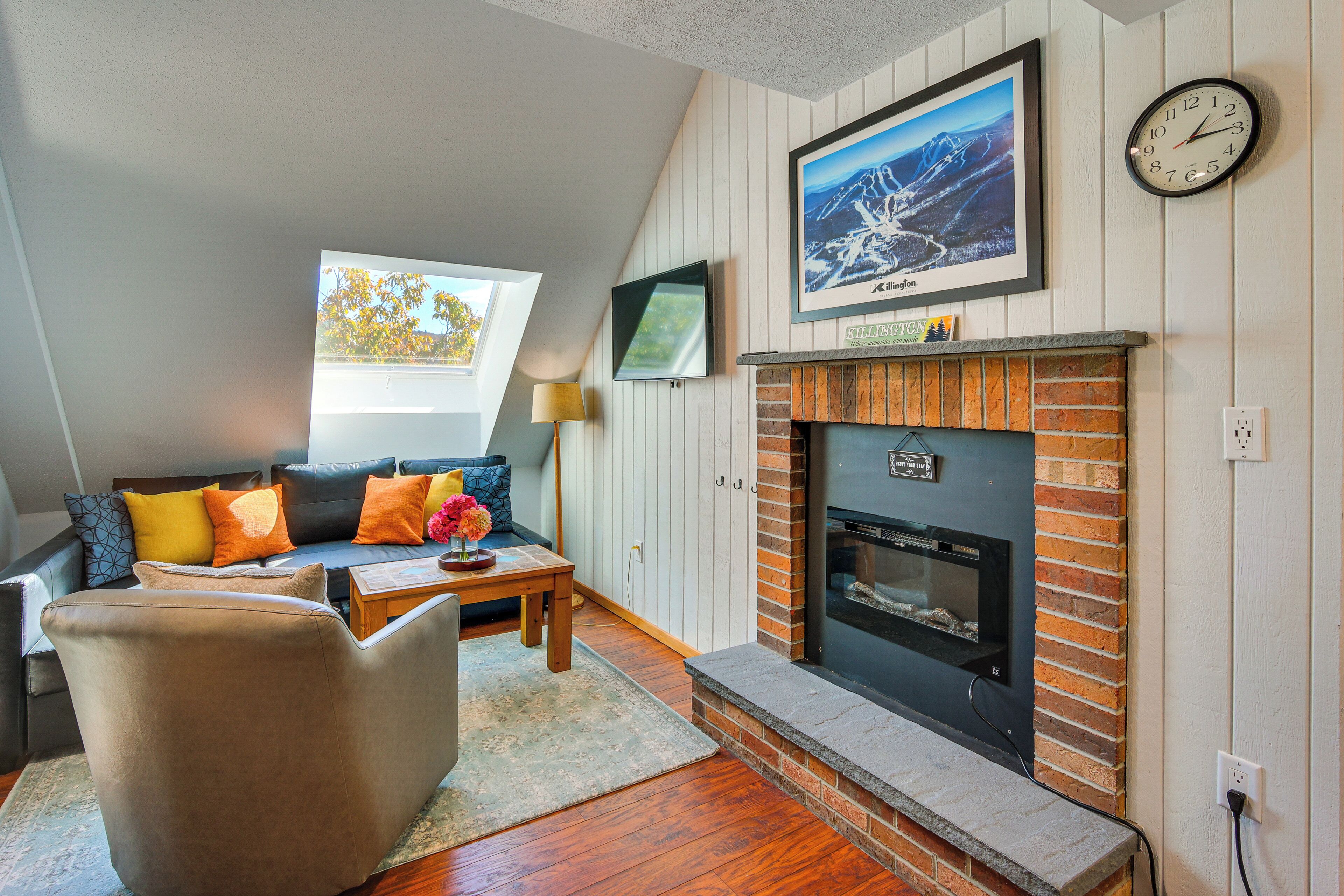Un salón acogedor con chimenea, un sofá con cojines de colores, un televisor montado en la pared y un reloj.