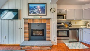 A modern kitchen with a brick fireplace, stainless steel appliances, and a mounted TV. - Steps to Ski Lifts! Mountain Green Resort Retreat (Killington)