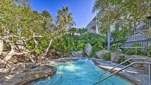 Ein Pool, umgeben von tropischer Vegetation und Felsen, mit wolkenlosem, blauem Himmel.