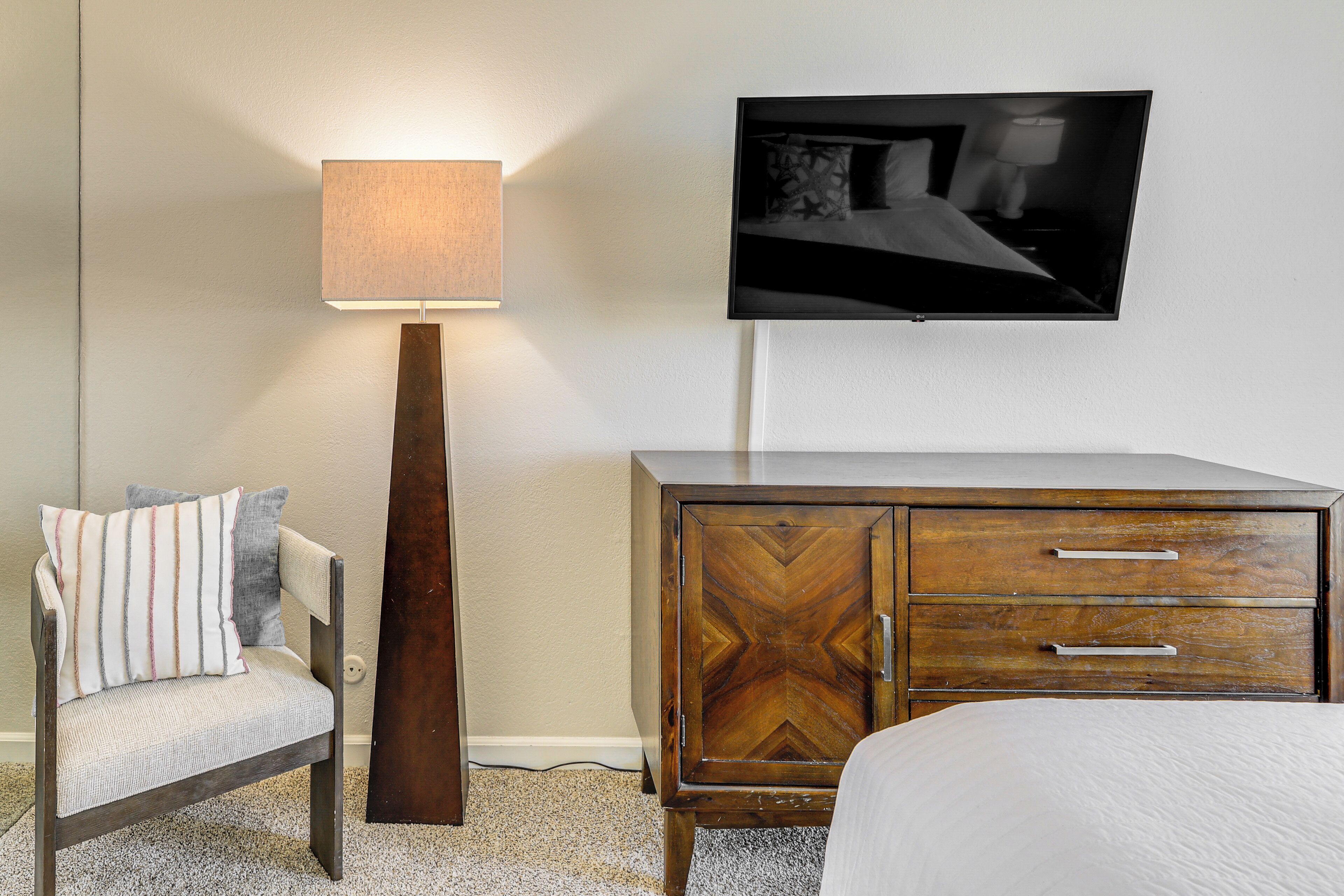 A hotel room with a bed, a wooden cabinet with drawers, a chair with a cushion, a floor lamp, and a flat-screen TV mounted on the wall.