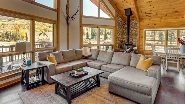 A spacious living room with a large sectional sofa, a coffee table, and a stone fireplace. Large windows offer a view of the surrounding forest. - Stunning San Juan Mtn Views: Cabin Near Skiing! (Durango)