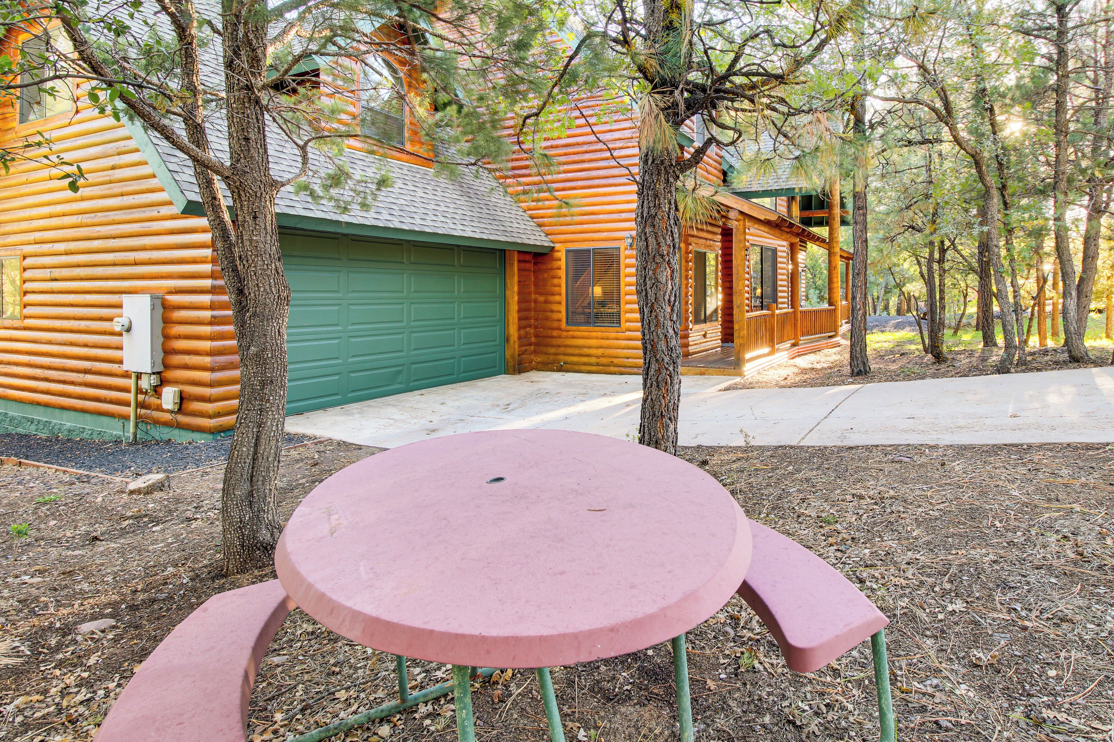 A log cabin with a green garage, a wooden porch, and a pink table in a wooded area.