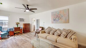 Una sala de estar con un sofá beige, una mesa de centro de vidrio, un sillón azul, un aparador de madera y un ventilador de techo.
