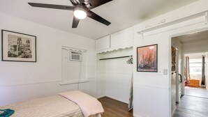 Un dormitorio con cama, ventilador de techo, ventana con aire acondicionado y puerta que da a otra habitación.