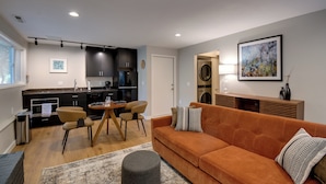 A modern living room with a sofa, a coffee table, and a dining area with chairs. There is a kitchen in the background with dark cabinets and a refrigerator. - Walk to Beach + Village: Bainbridge Island Gem! (Bainbridge Island)