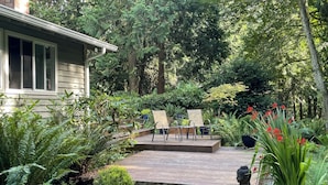 A wooden deck with two chairs and a table, surrounded by lush greenery and a variety of plants. - Walk to Beach + Village: Bainbridge Island Gem! (Bainbridge Island)